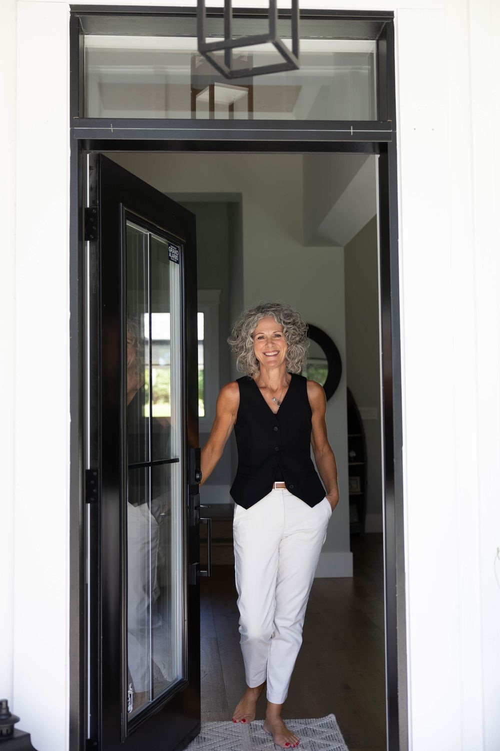 Smiling mature woman with gray hair standing in open doorway wearing black vest and white pants.