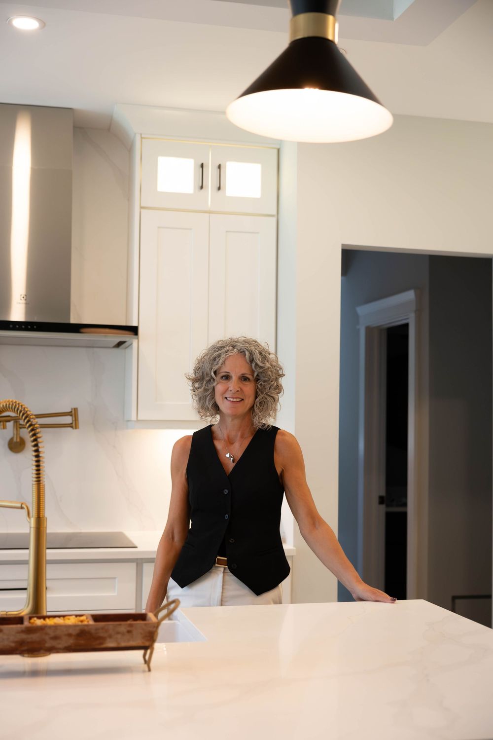 A smiling woman with curly gray hair stands in a modern white kitchen with gold fixtures and a marble island.