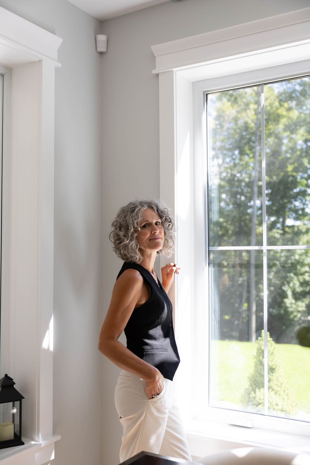 Elegant mature woman with silver curly hair in black sleeveless top standing by bright window in modern home.