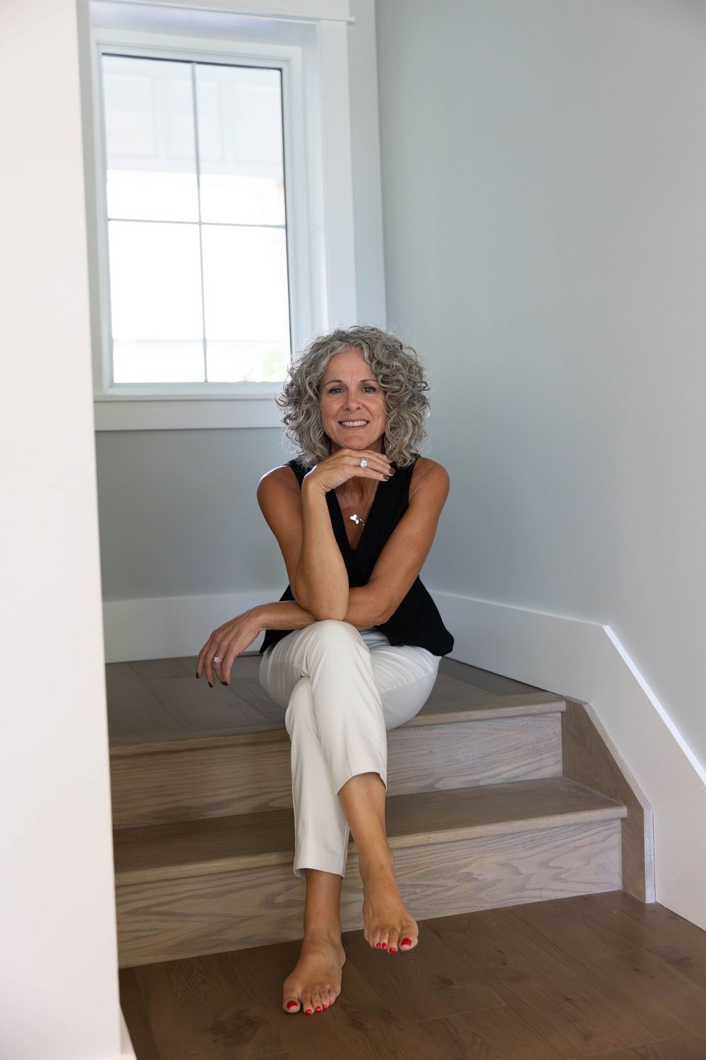 Smiling middle-aged woman with gray curly hair sitting barefoot on wooden stairs in a bright home interior.