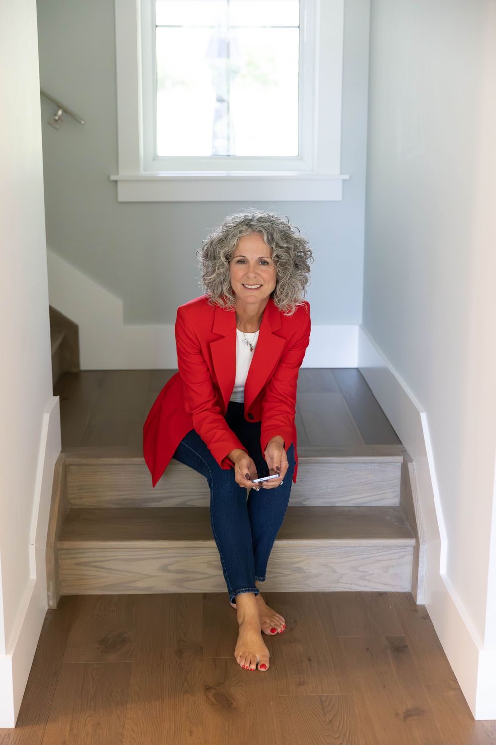Smiling woman with gray curly hair in red blazer and jeans sits barefoot on wooden stairs holding a phone.