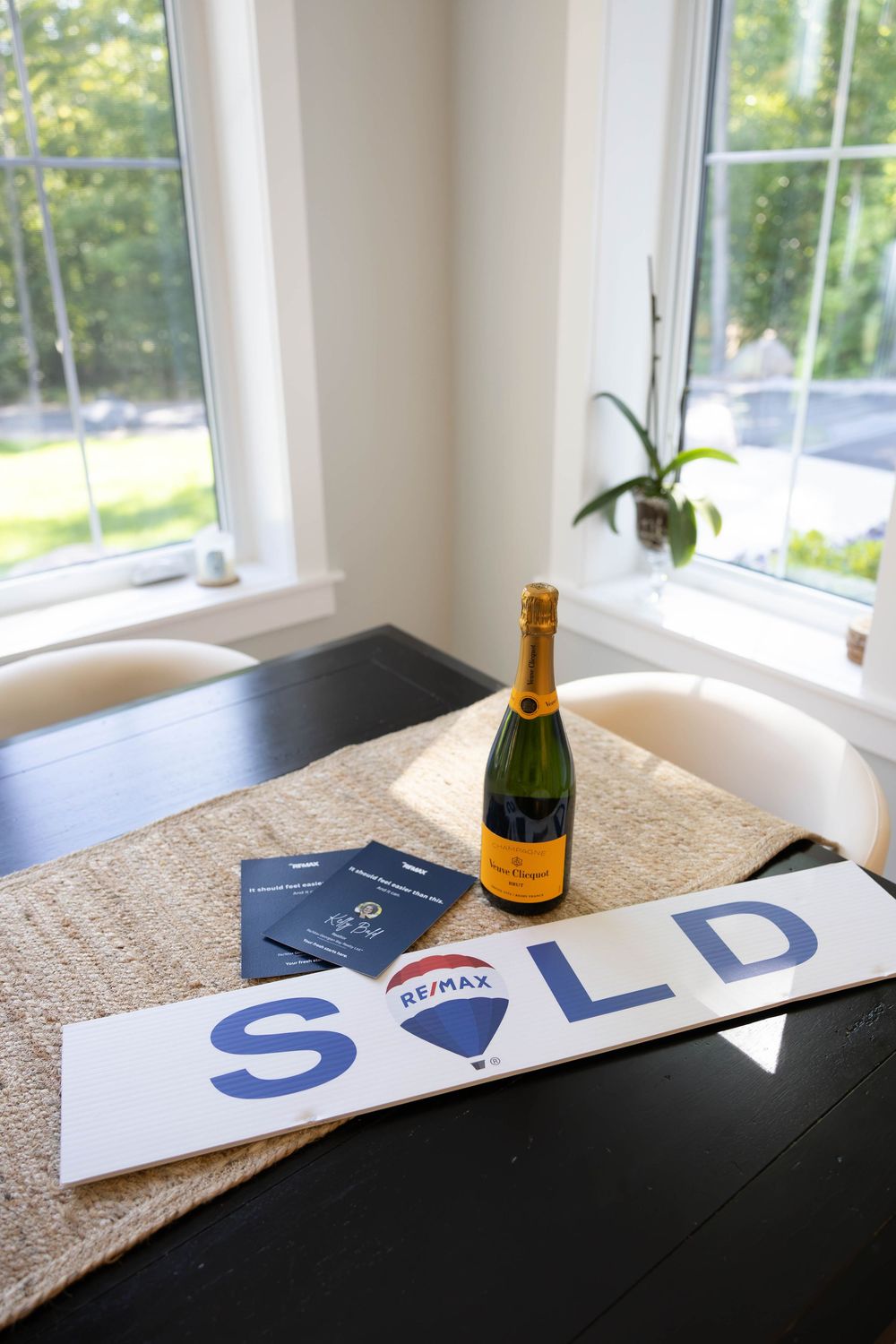 RE/MAX SOLD sign with Veuve Clicquot champagne bottle on dark table celebrating a home sale closing.