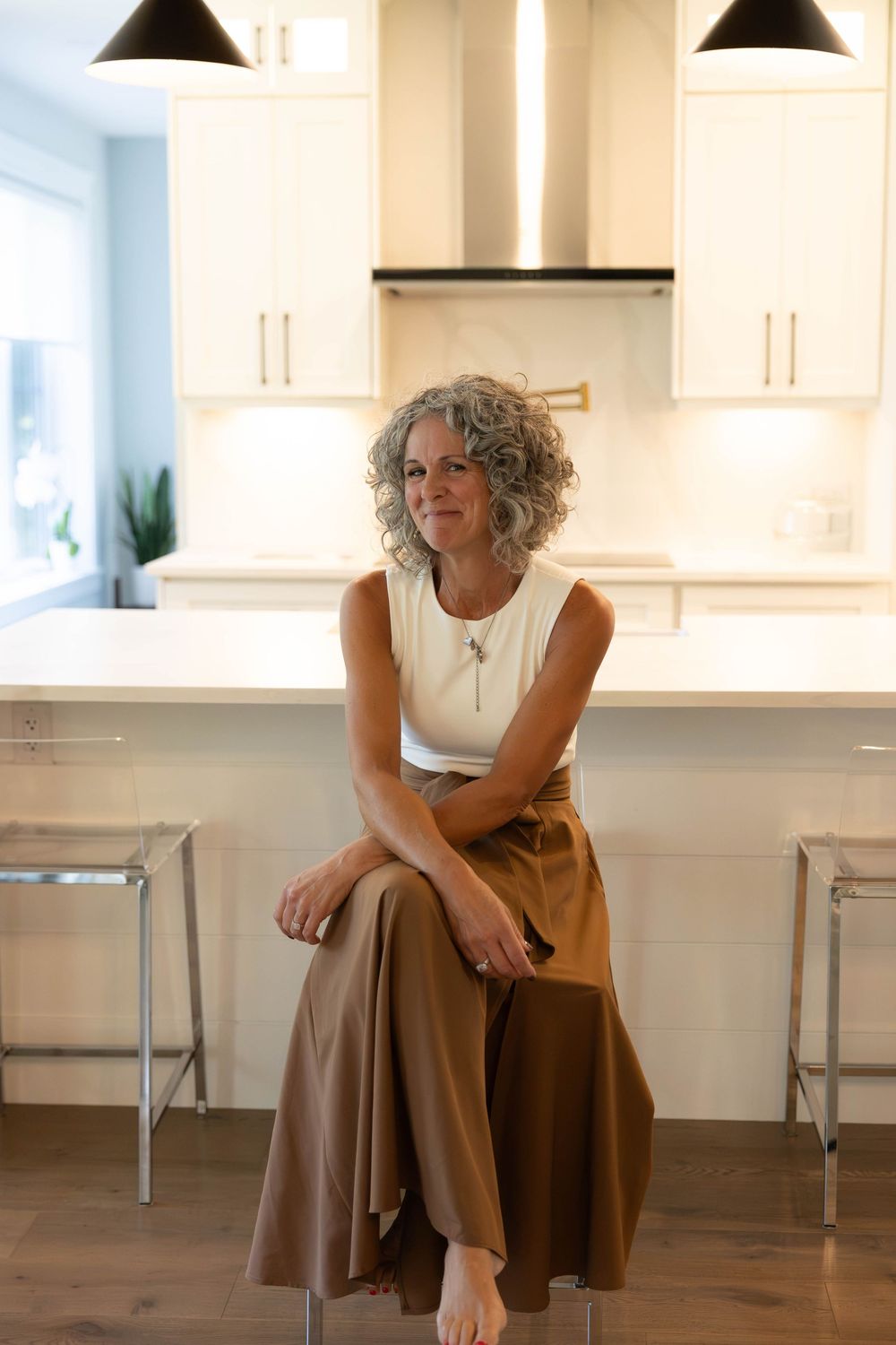 A woman with curly gray hair sits on a barstool in a modern white kitchen, wearing a white top and brown skirt.
