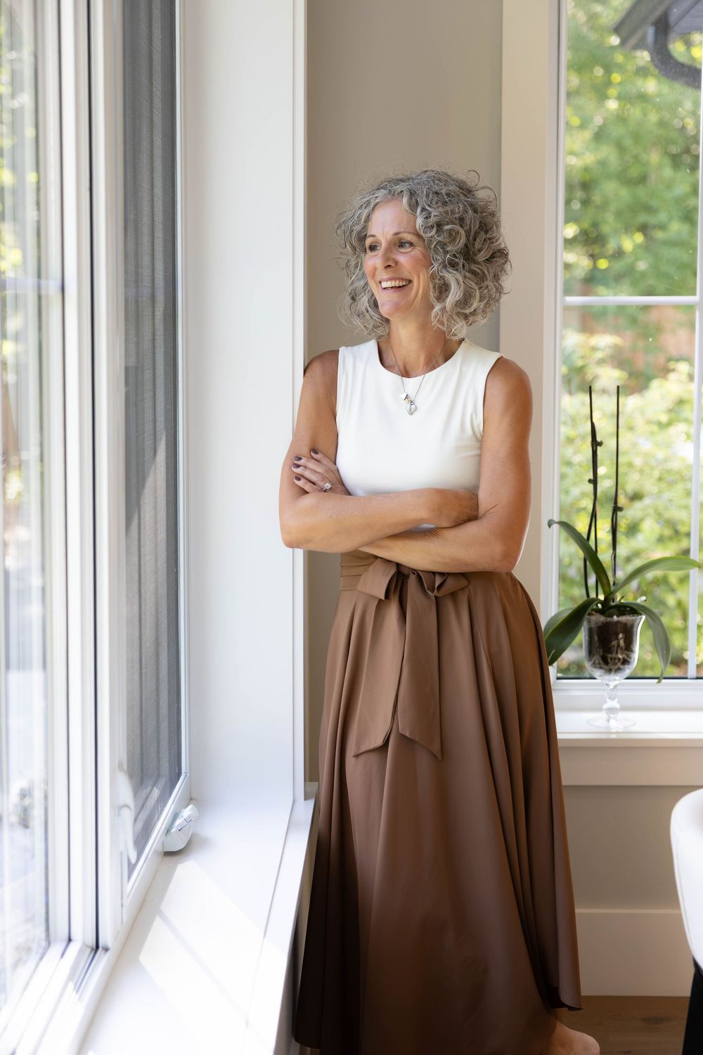 Elegant older woman with gray curly hair in white top and brown skirt stands by window with arms crossed, smiling.
