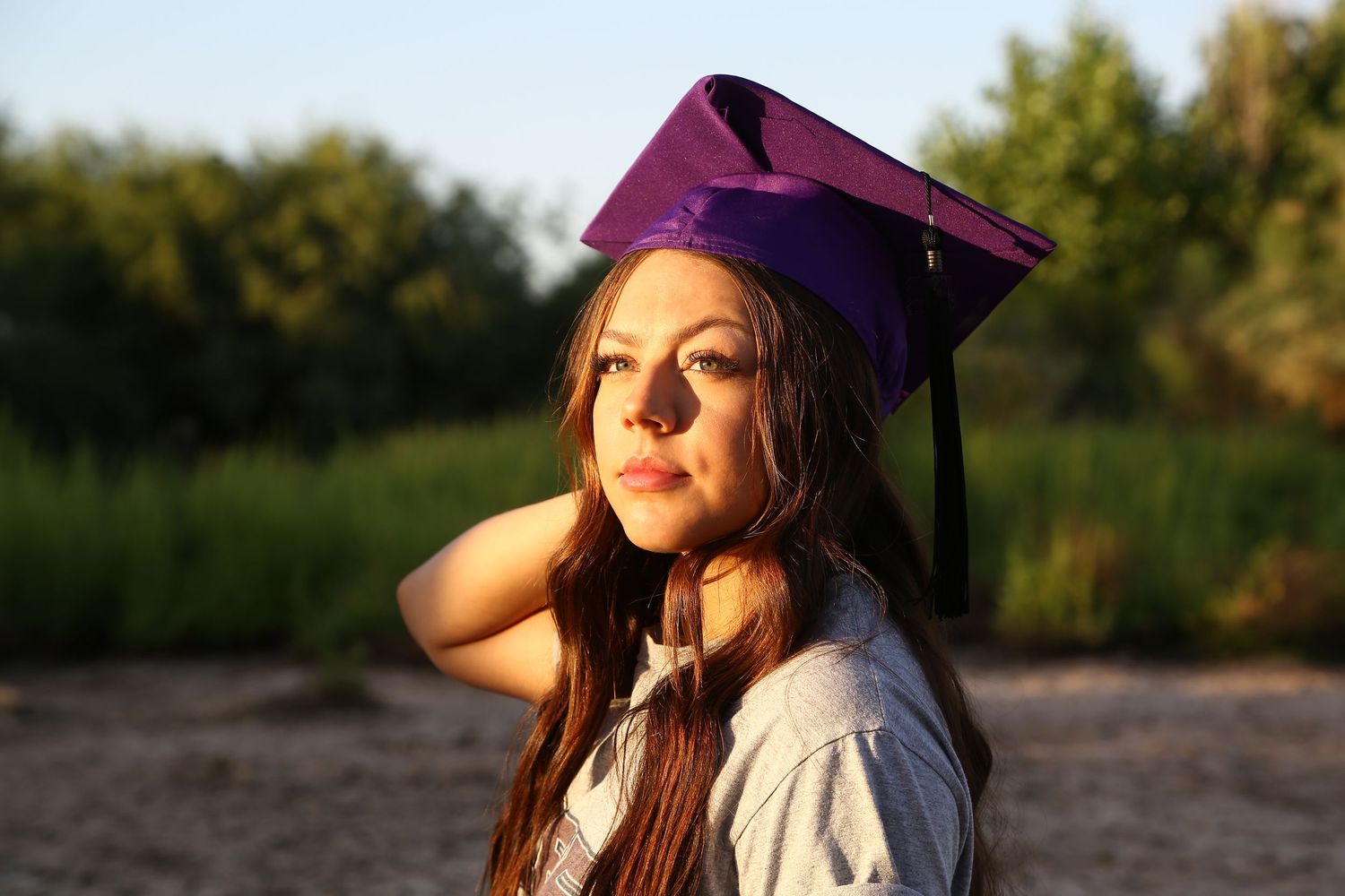 Young woman in a purple graduation cap with a black tassel poses outdoors in warm golden sunlight with green foliage behind.