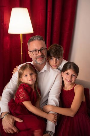A man with glasses hugs three children in red outfits, posing warmly in front of red curtains with a lamp.