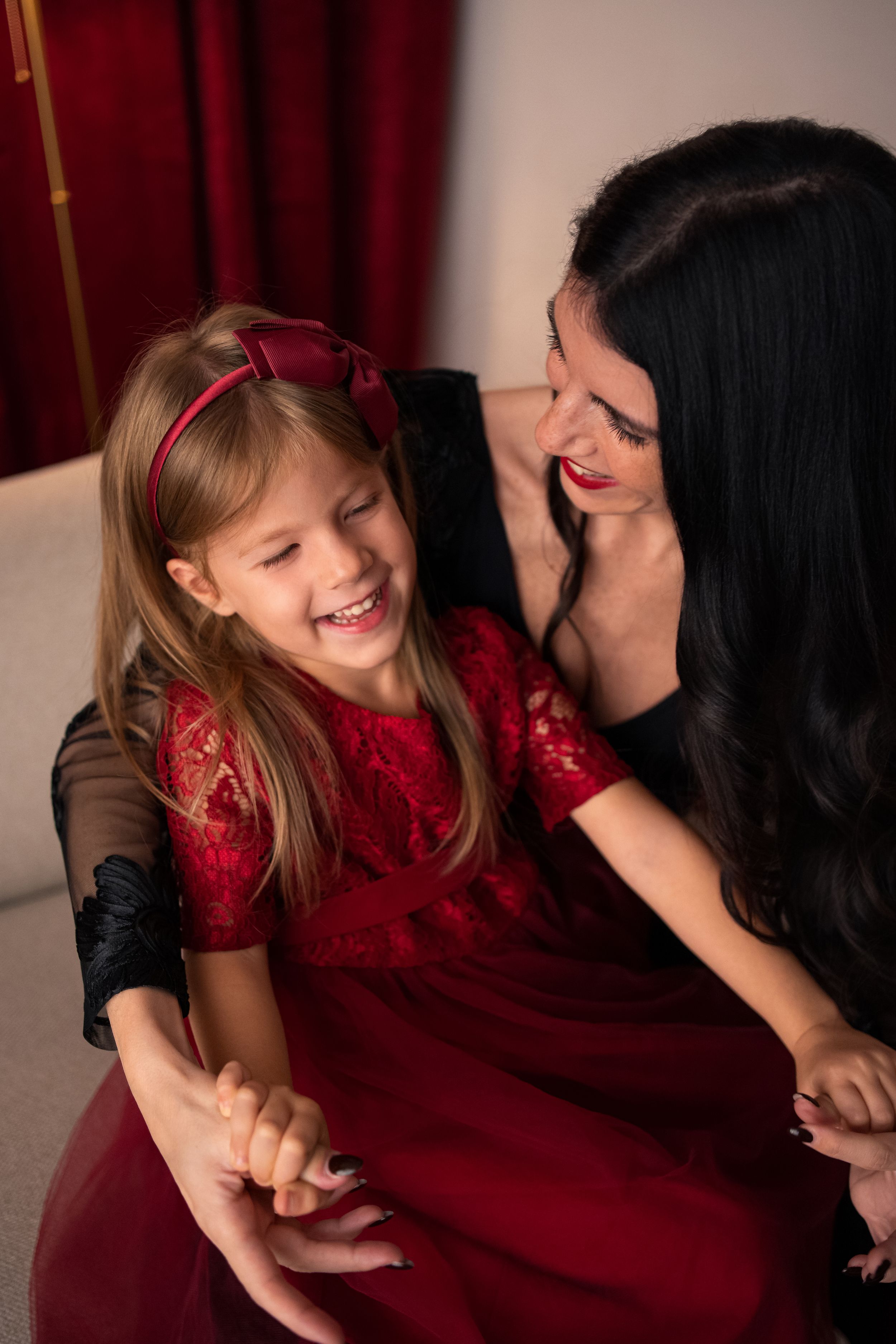 A laughing little girl in a red dress shares a joyful moment with a dark-haired woman in matching red attire.
