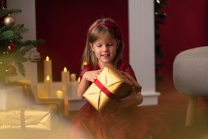 Young girl in red dress smiling while holding a gold Christmas gift box near a decorated holiday tree.