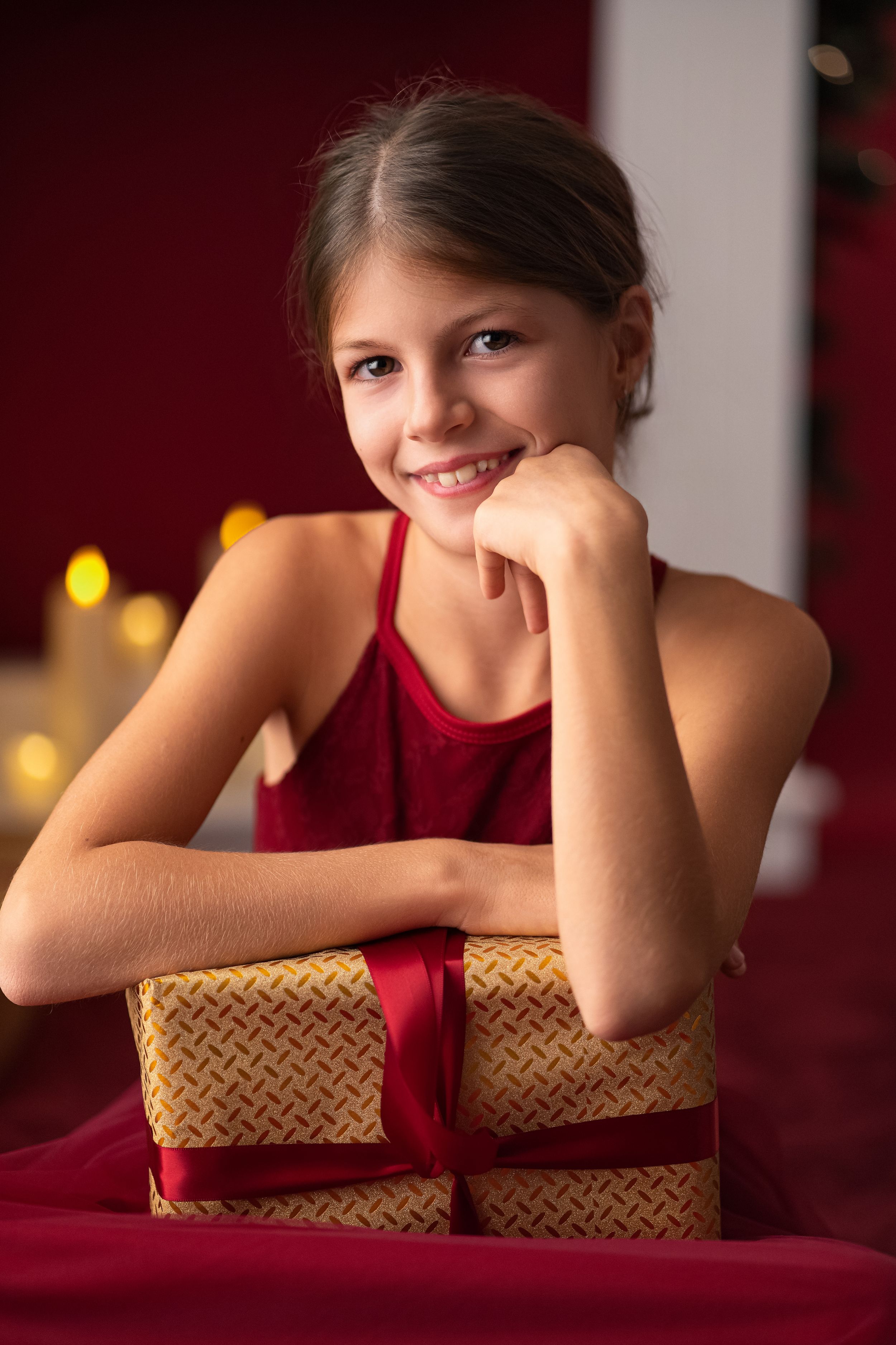 Smiling young girl in red dress leaning on a gold gift box with red ribbon in a festive holiday setting.