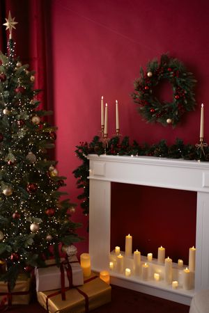 Festive Christmas room with decorated tree, white fireplace filled with candles, wreath, and gifts against a red wall.