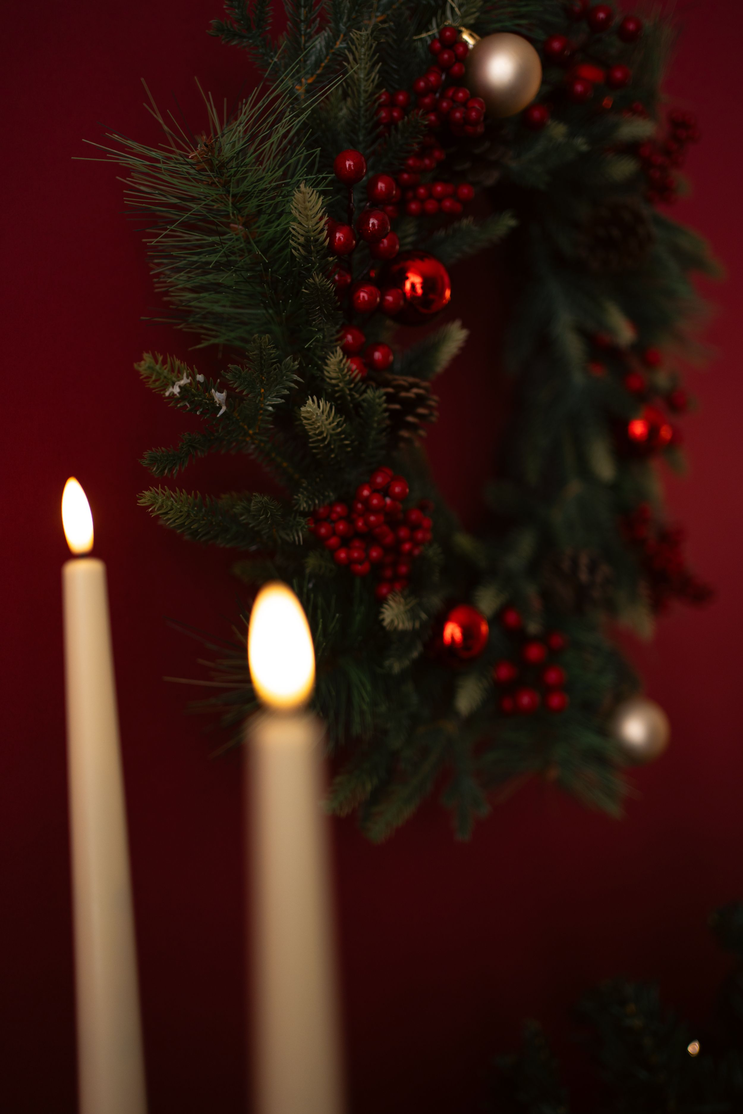 Christmas wreath with red berries, pine cones, and ornaments on red background with two lit white candles.