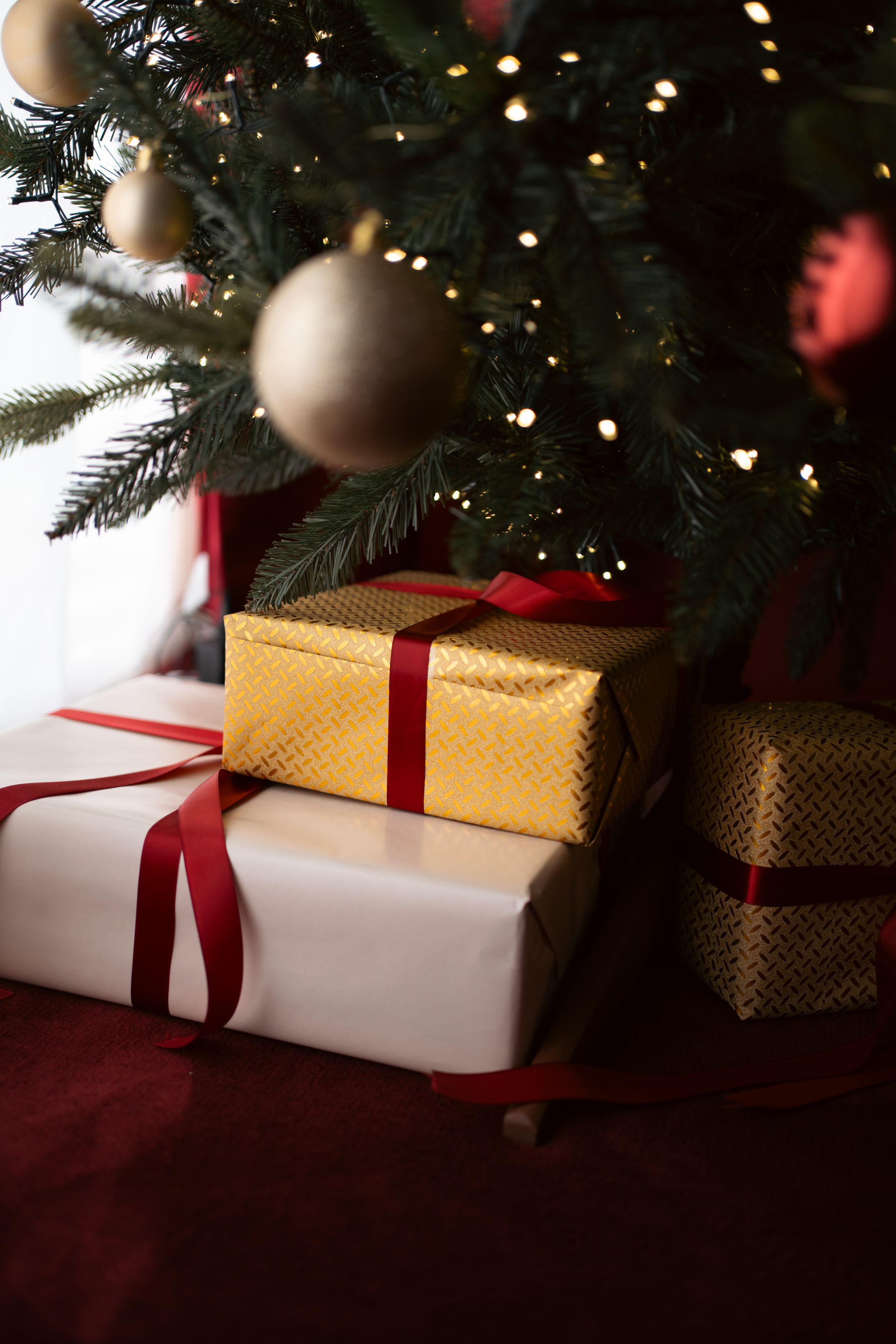 Wrapped Christmas gifts with red ribbons placed under a decorated tree with gold ornaments and warm glowing lights.