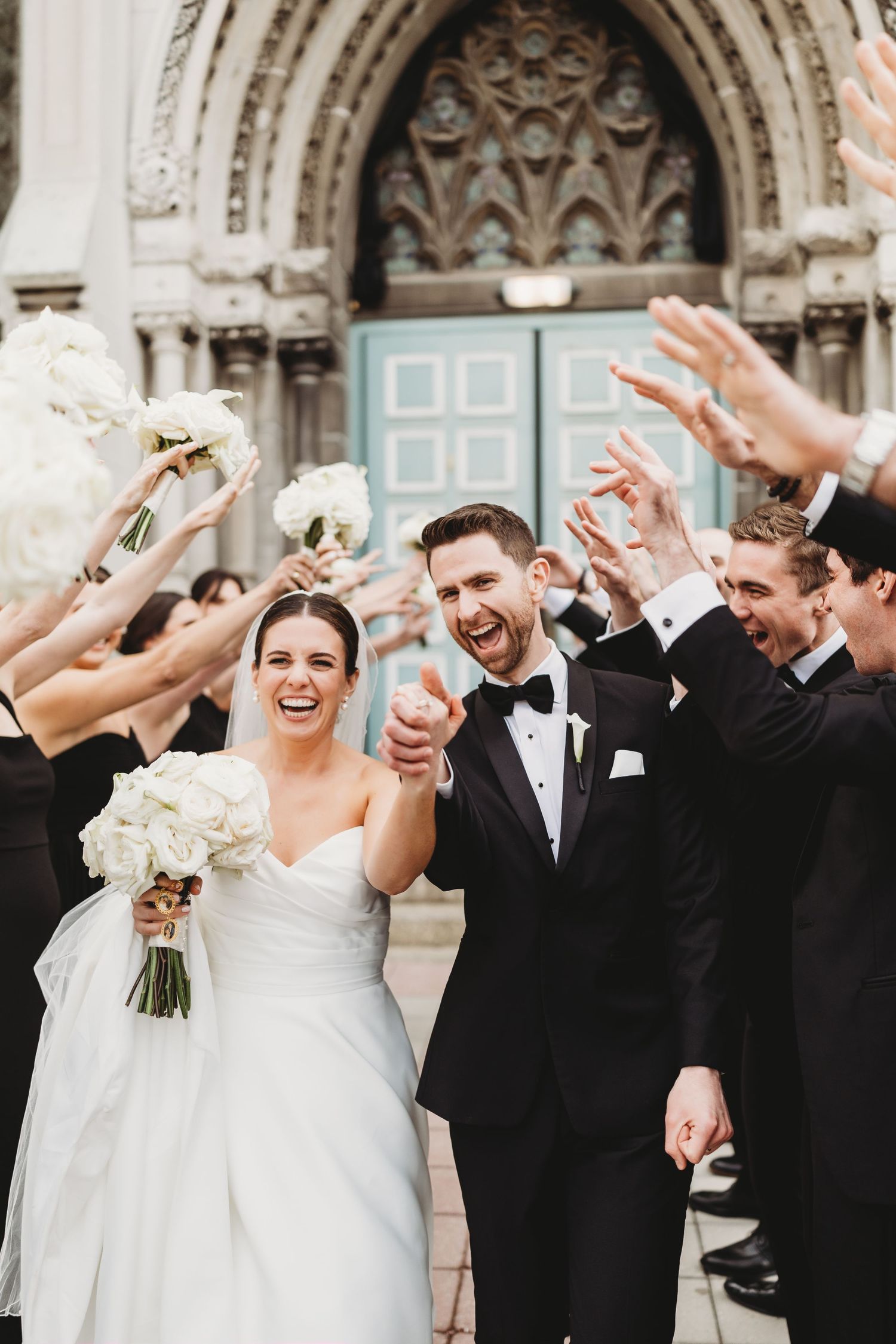 A smiling bride in a strapless white gown holds a bouquet while a groom in a black tuxedo embraces her outdoors.