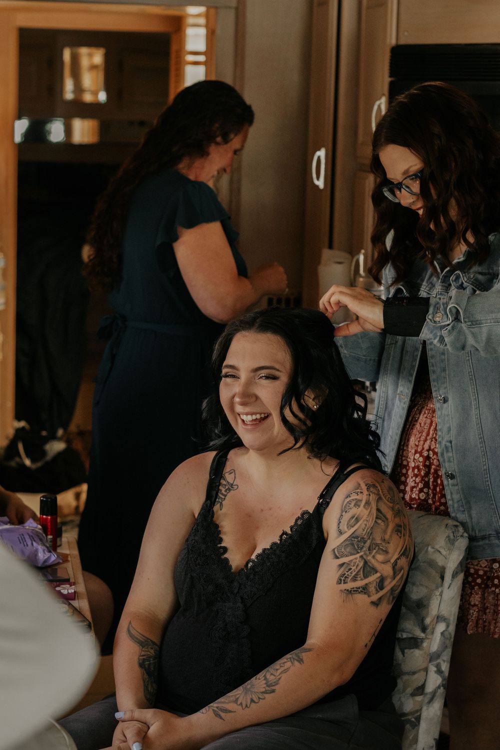 Woman laughing while getting hair styled by makeup artist, with tattoos visible, during wedding preparations indoors.
