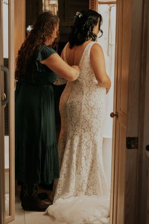 Bride in white lace mermaid gown being helped by bridesmaid in green dress in a doorway.