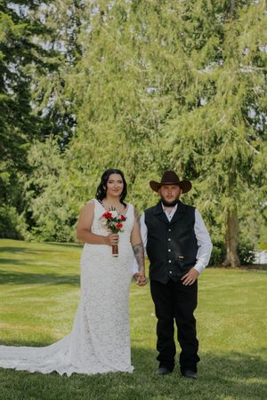 Bride in lace gown holding red roses stands beside groom in cowboy hat and vest on green lawn.