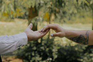 Two hands gently touching outdoors, one with a tattoo, sharing a tender moment in a lush green garden setting.