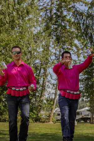 Two men in matching pink shirts with beer can belts walking outdoors at a wedding celebration.