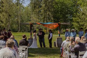 Outdoor wedding ceremony under colorful orange and blue fabric arch with guests seated on white chairs in lush green setting.