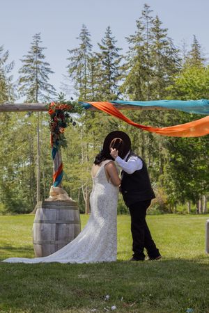 Bride and groom share first kiss under colorful draped arch at outdoor wedding ceremony surrounded by tall evergreen trees.