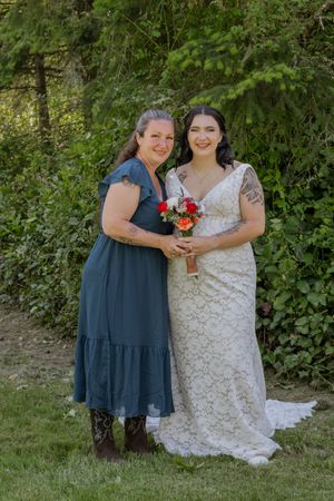 Bride in lace gown holds bouquet beside older woman in blue dress, posing outdoors among green trees.
