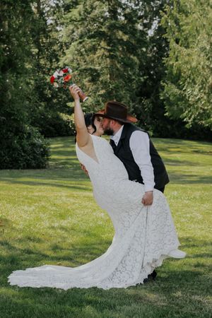 Bride in lace gown holds bouquet aloft as groom in cowboy hat dips her for a romantic kiss on green lawn.