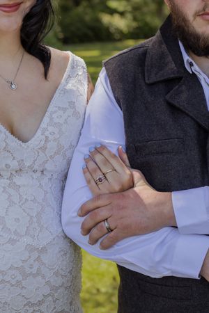 Bride and groom linking arms, showcasing wedding rings, bride in lace dress with necklace, groom in gray vest.