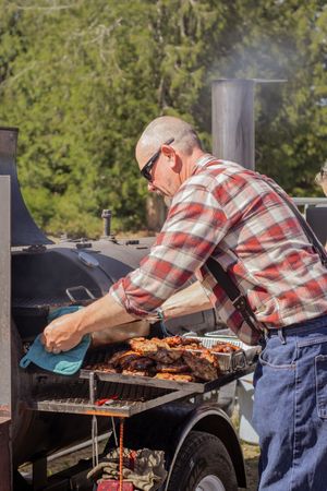 Man in plaid shirt grilling BBQ meats on large smoker grill outdoors on sunny day.