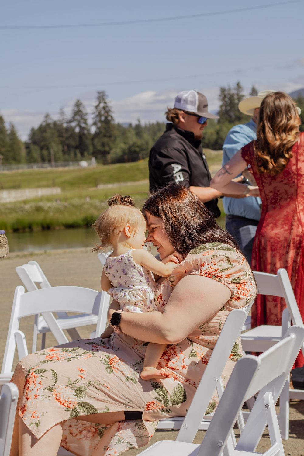Woman in floral dress holding a baby at an outdoor wedding ceremony with white chairs near a scenic river.