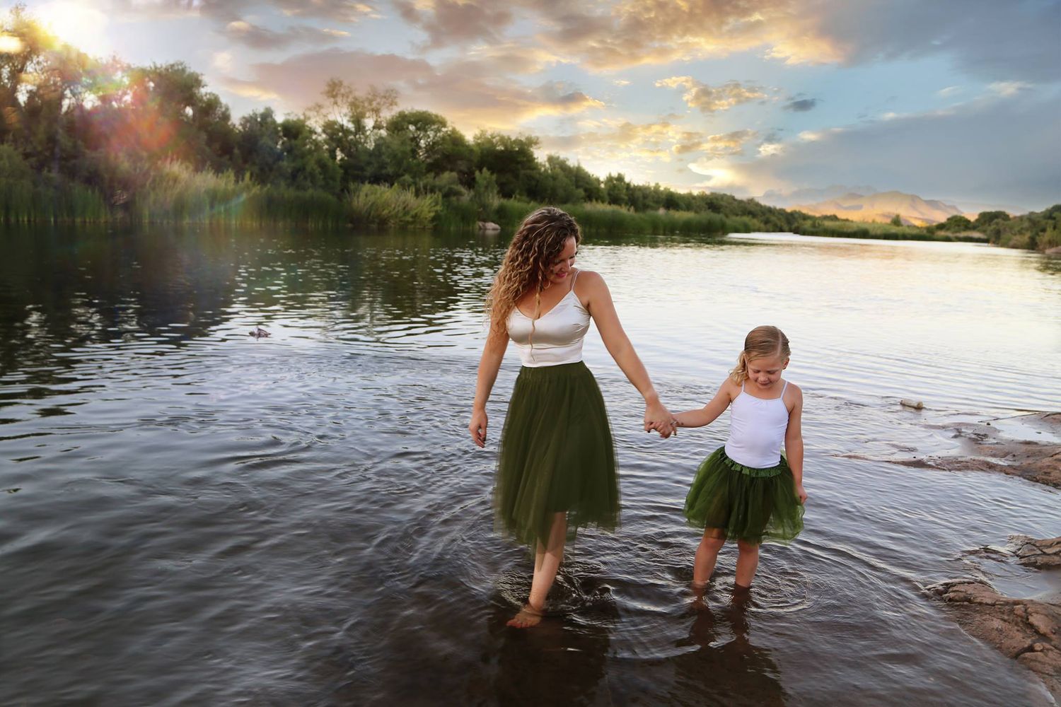 A woman and a young girl in green tulle skirts hold hands while wading in a calm river at golden sunset.
