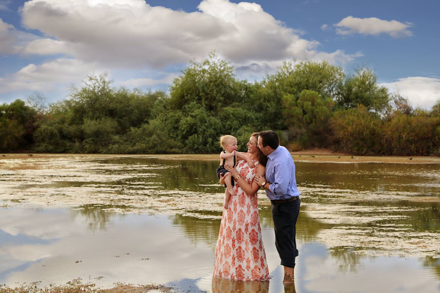 A couple holds their baby while standing in shallow water at a lake with green trees reflected under a bright sky.