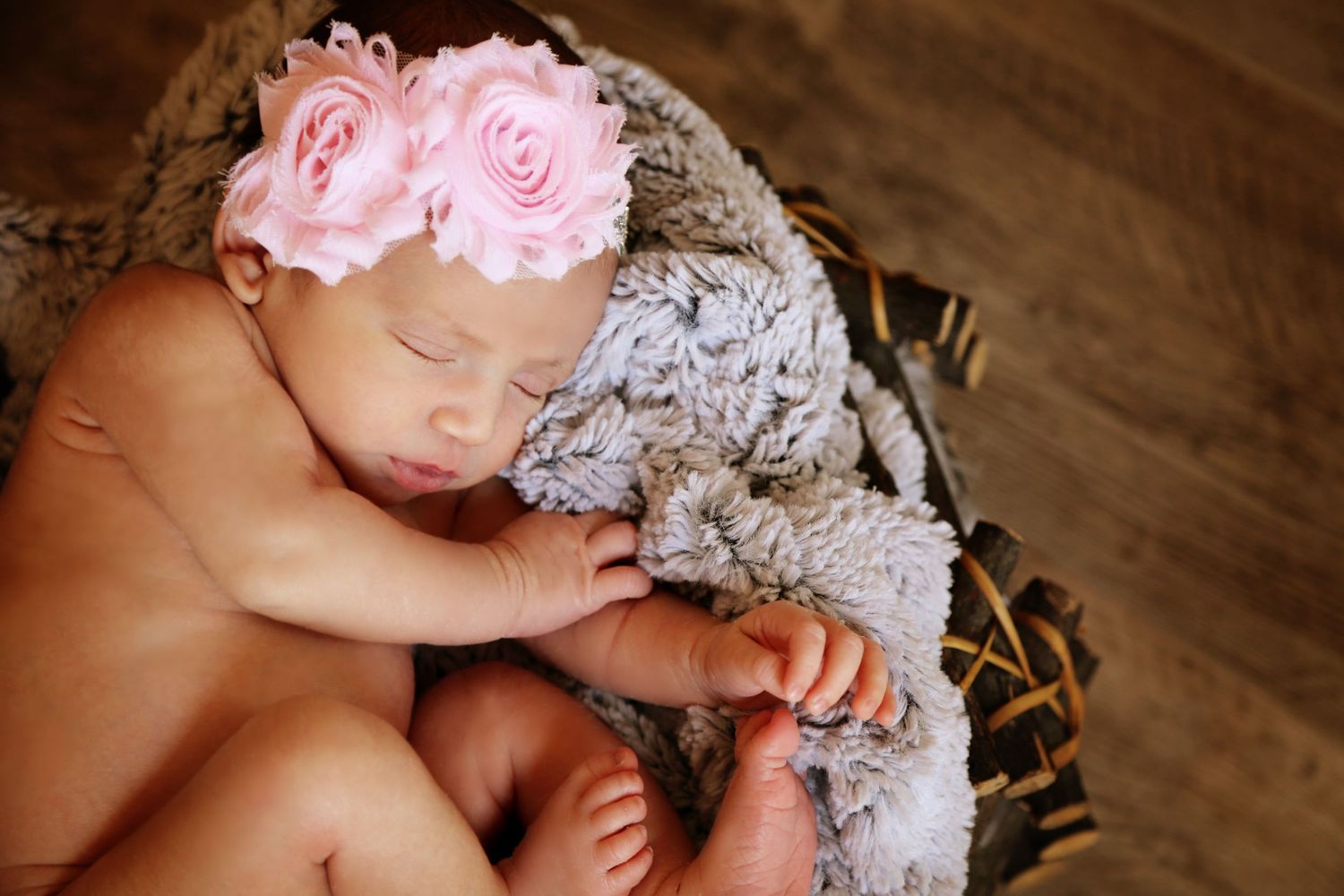 A sleeping newborn baby wearing a pink flower headband rests in a basket lined with soft gray fur on a wooden floor.