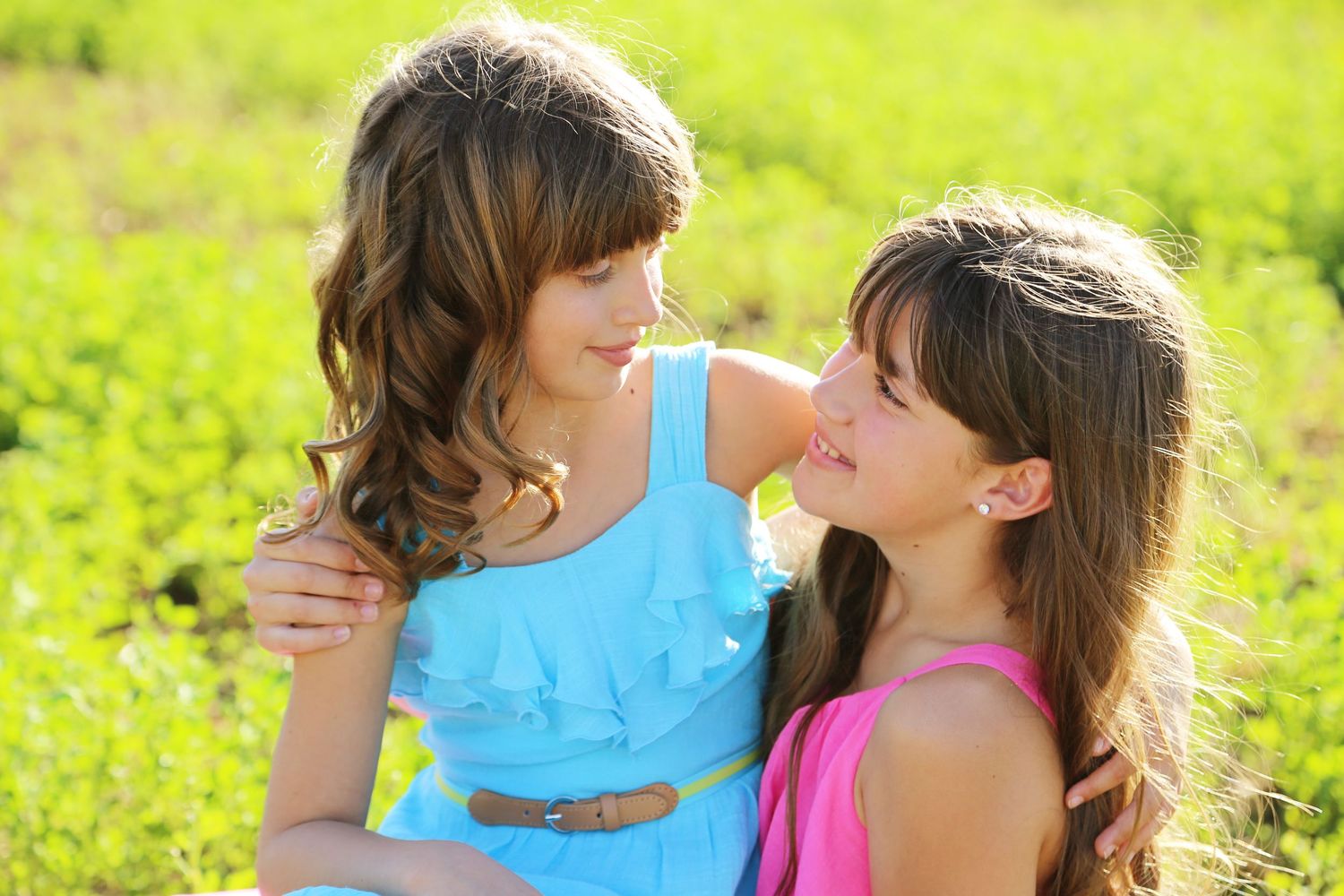 Two smiling girls in blue and pink dresses embrace and look at each other in a sunny green field.