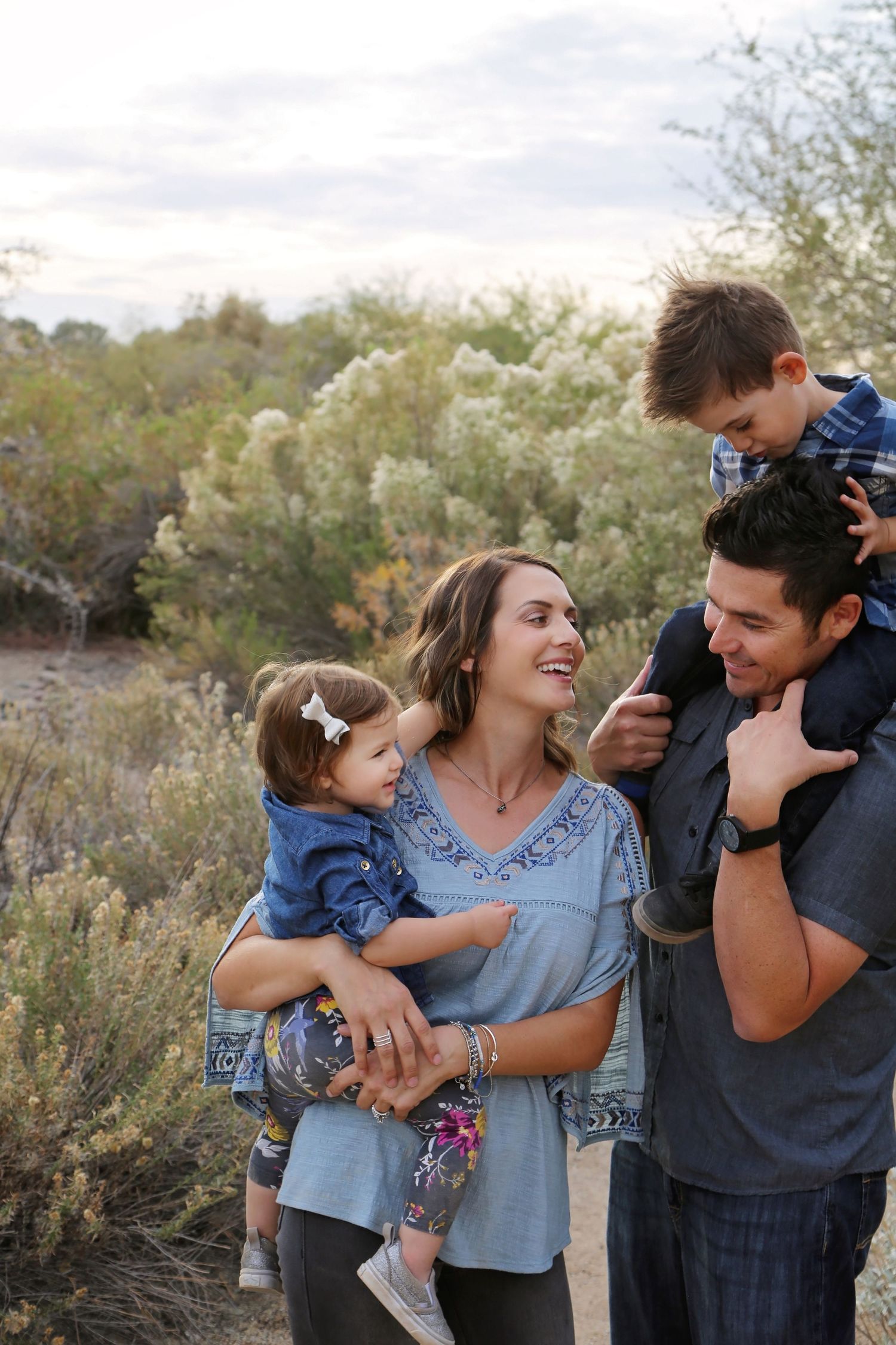 A smiling mother holds her baby daughter while a father carries their young son on his shoulders in a desert landscape.