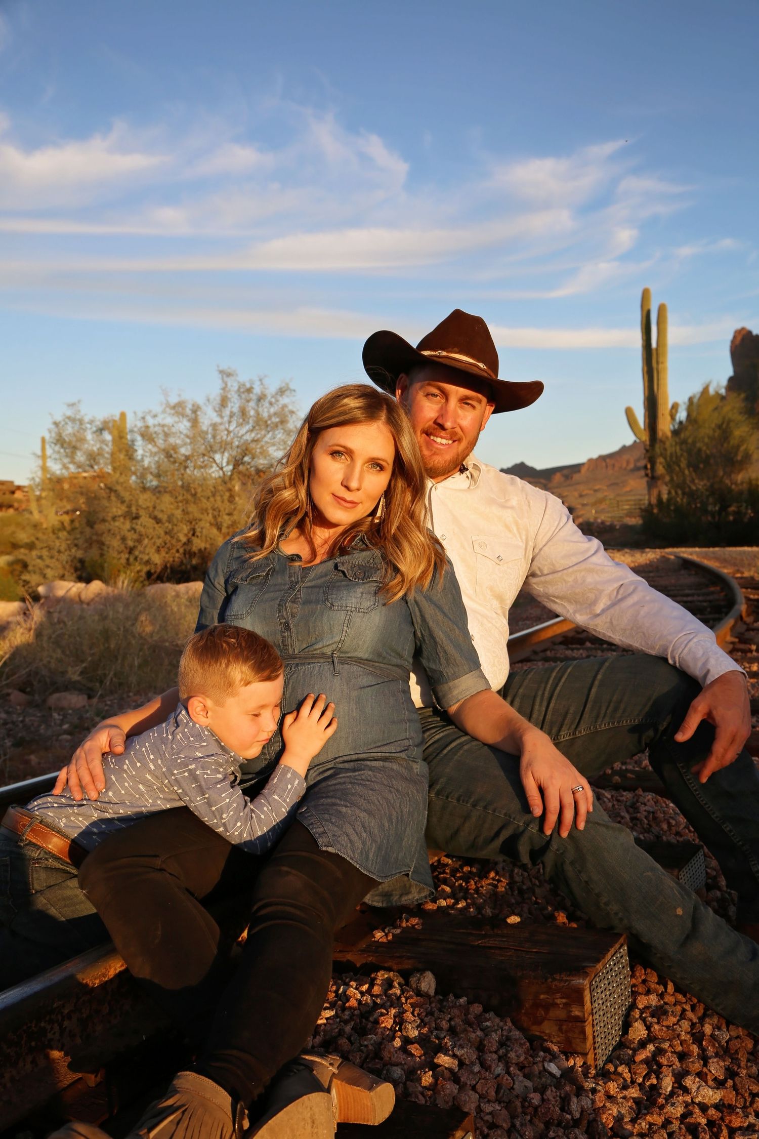 An expectant mother in a denim shirt sits with her young son and cowboy hat-wearing husband on railroad tracks in the desert.