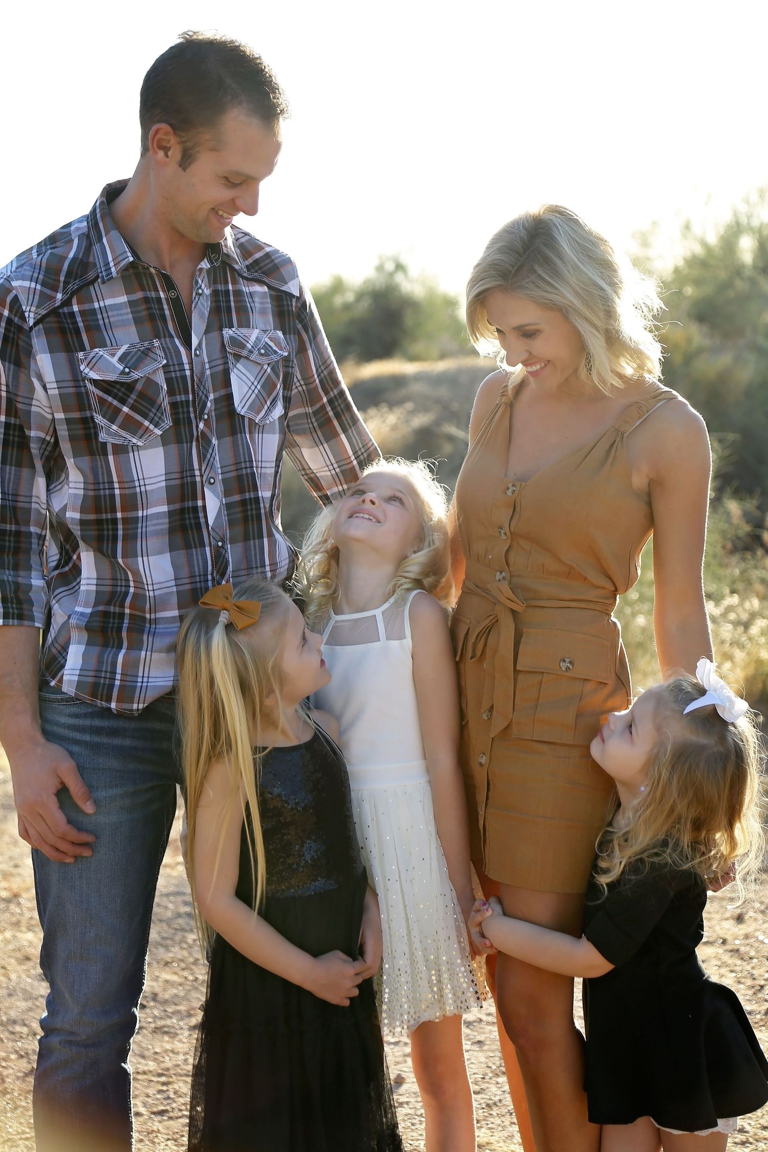 Smiling parents stand with their three young daughters in matching dressy outfits outdoors in warm golden sunlight.