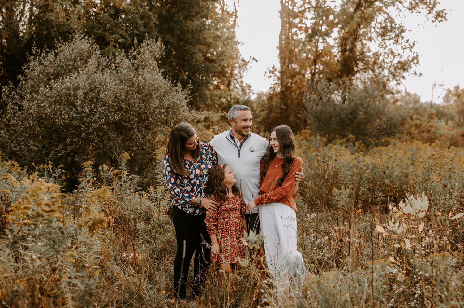 Family of four smiling and laughing together in a golden autumn meadow surrounded by lush greenery.