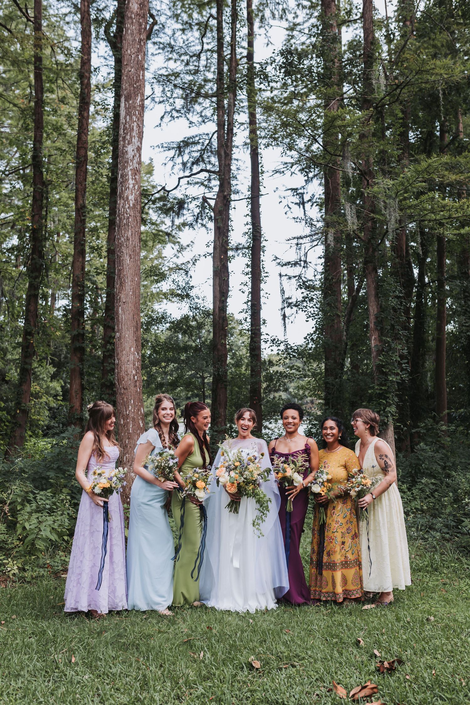 A bride and her seven bridesmaids in colorful mismatched dresses hold bouquets while posing in a lush forest setting.