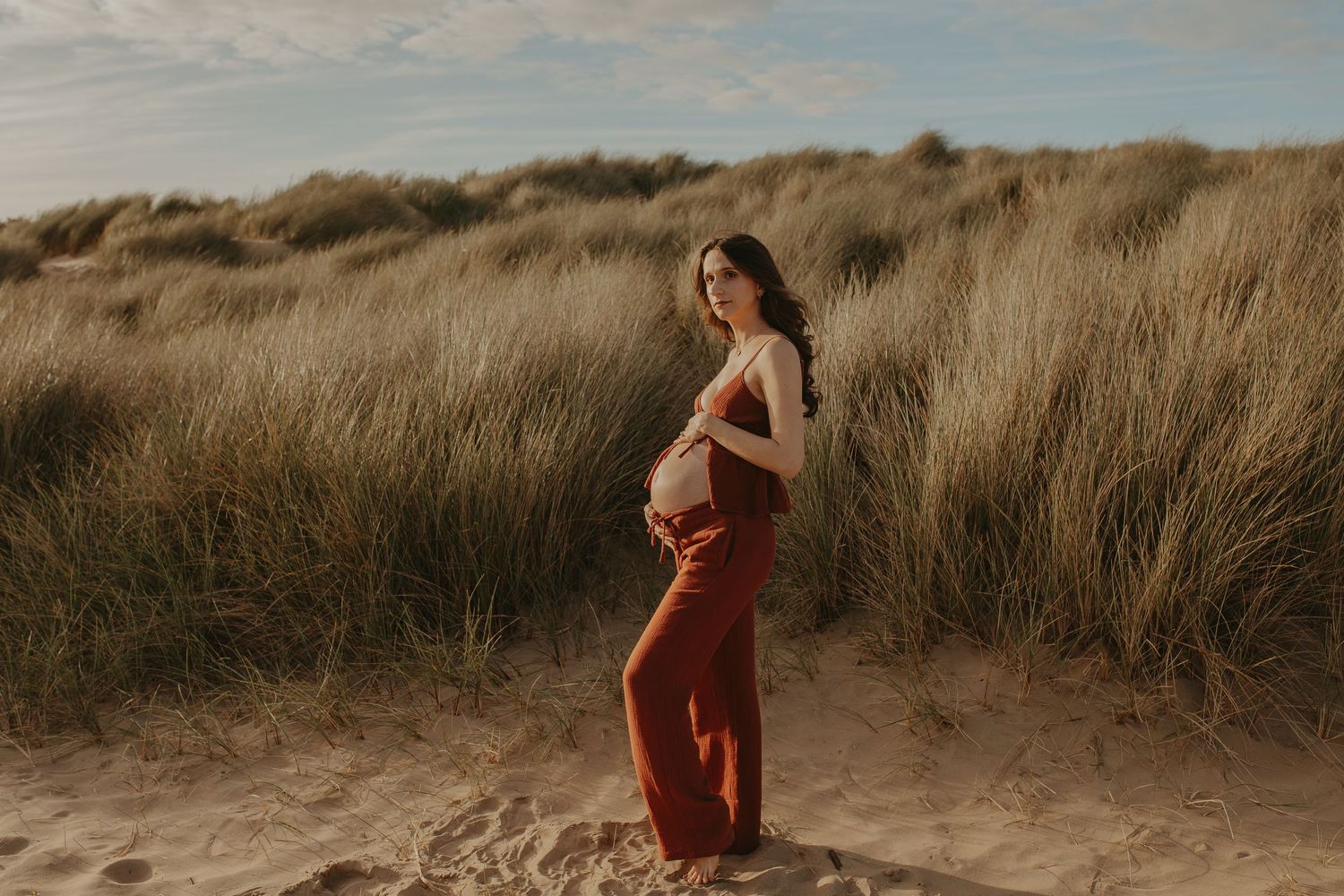 Pregnant woman in rust pants cradles baby bump on sandy beach dunes with golden grass backdrop.