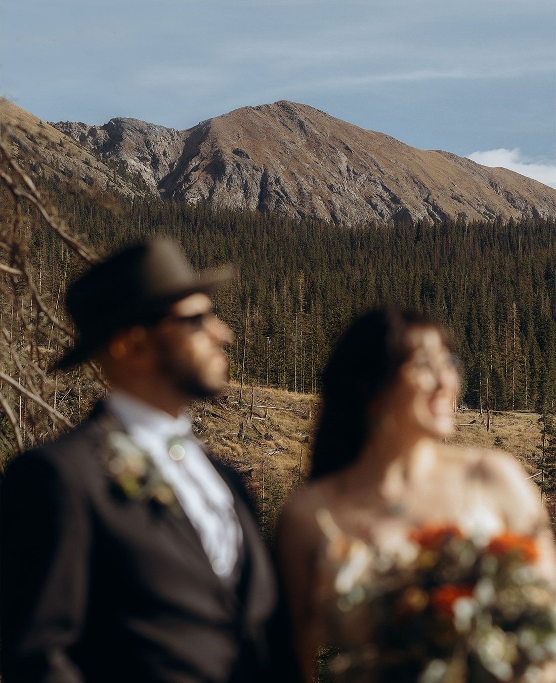 A blurred couple in wedding attire poses before a stunning mountain forest backdrop under a clear blue sky.
