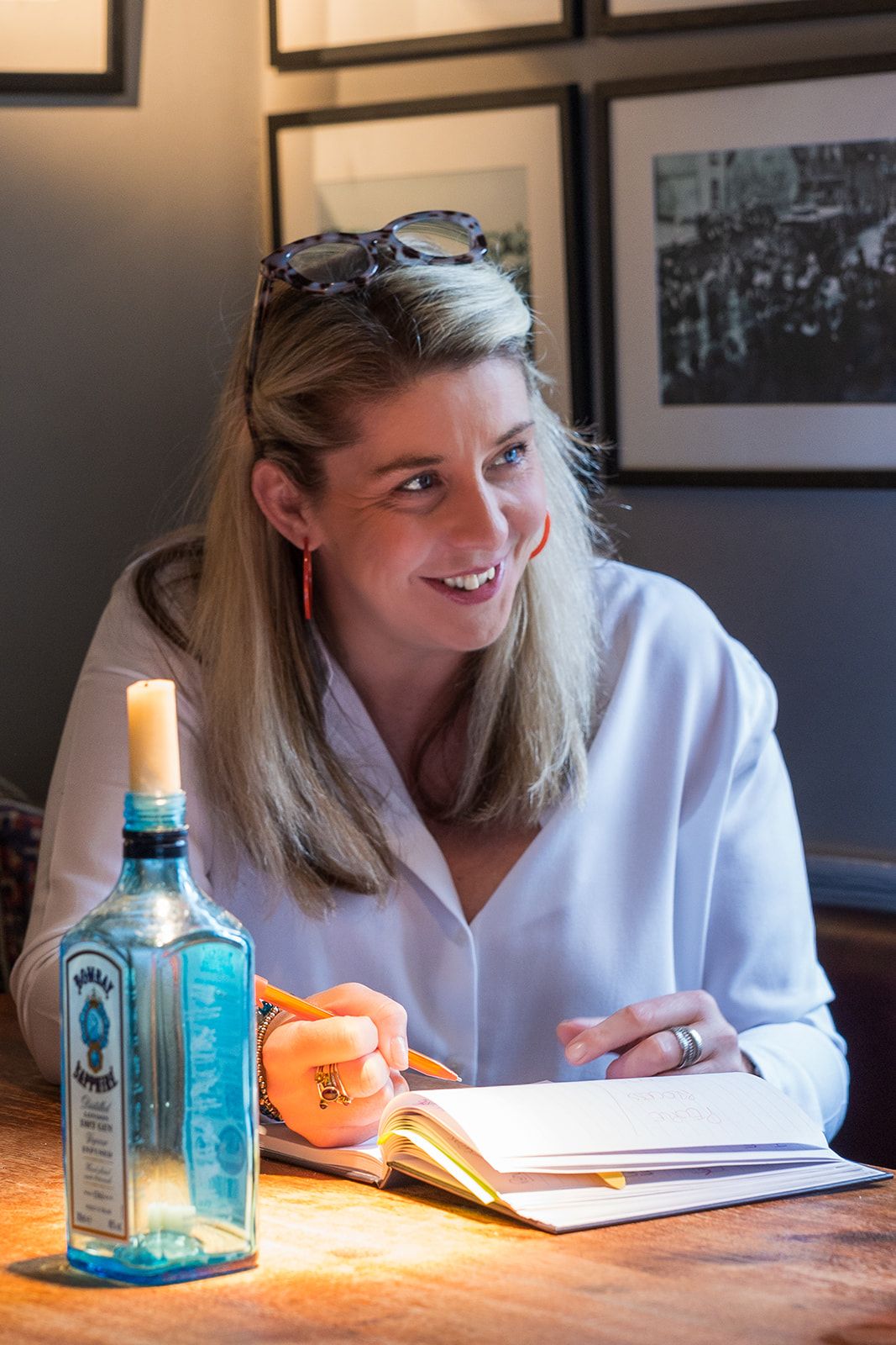 Smiling woman writing in notebook at table with Bombay Sapphire bottle in warm ambient light.
