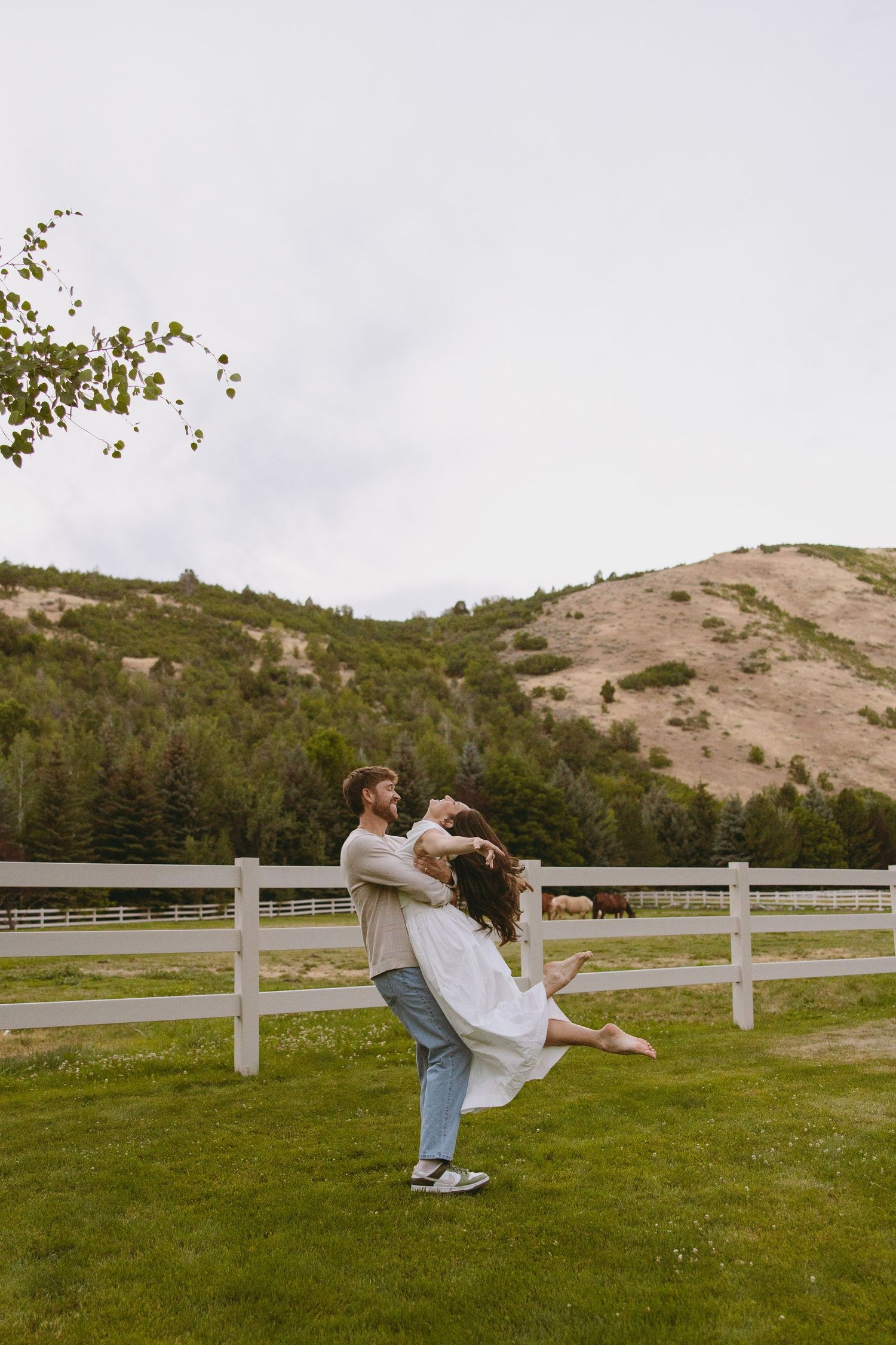 Bride in flowing white dress holds bouquet while standing on wooden deck surrounded by evergreen mountains at sunset.