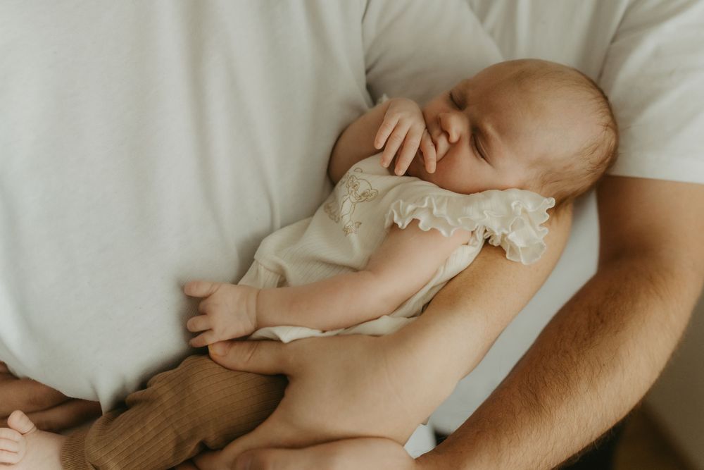 A newborn baby sleeping peacefully in a parent's arms, dressed in a delicate cream lace romper.