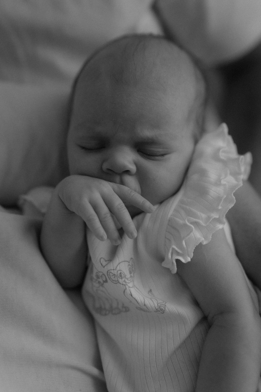 Black and white photo of a sleeping newborn baby in a white ruffled outfit, gently sucking on tiny fingers.