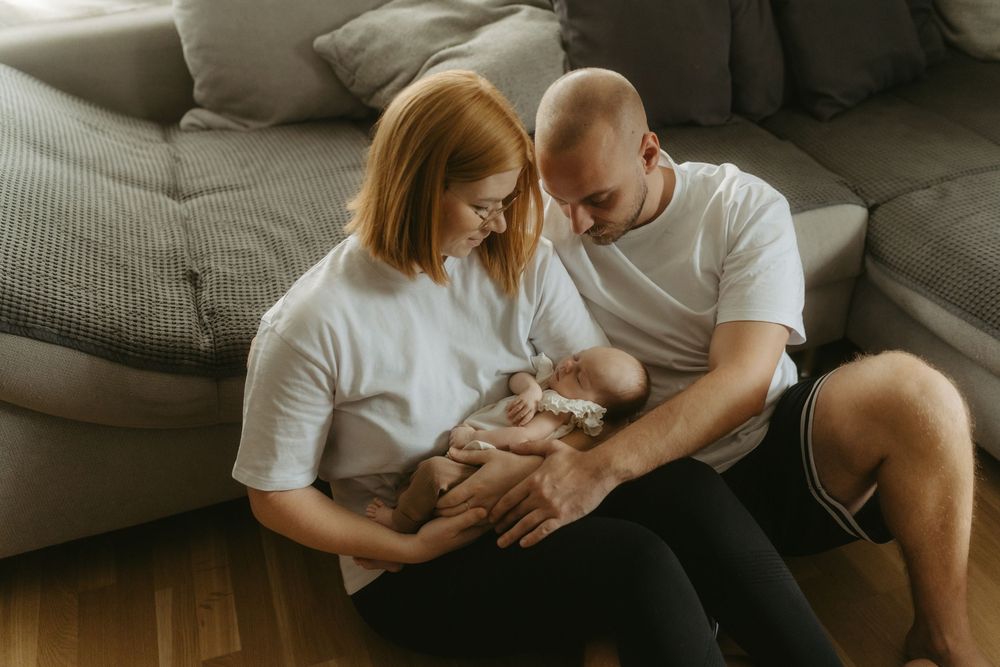 A couple in white t-shirts sit on the floor cradling their newborn baby together near a gray sofa.