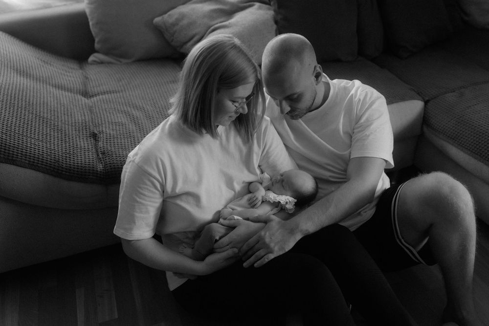 Black and white photo of parents lovingly cradling their newborn baby together on a couch at home.