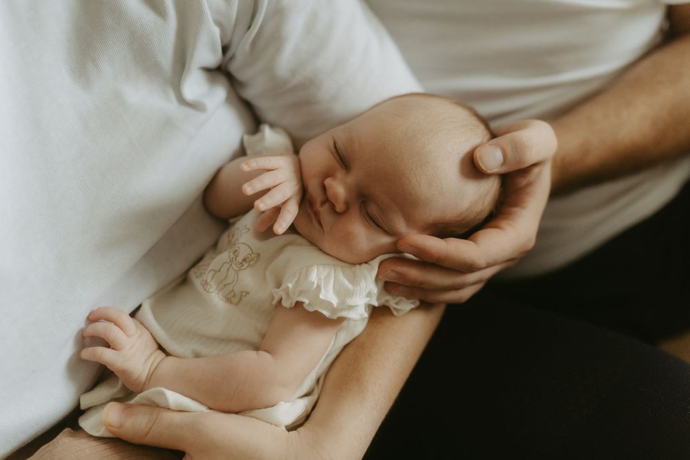 A newborn baby sleeping peacefully in a parent's gentle hands, wearing a white outfit.