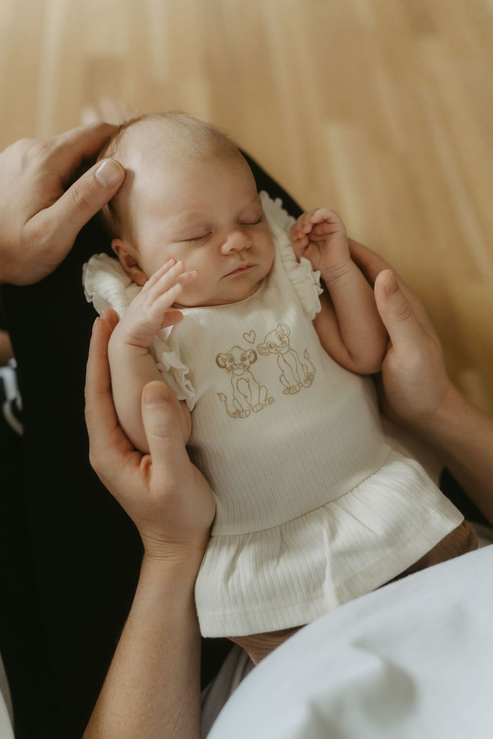 Newborn baby sleeping peacefully in parents hands, wearing a white embroidered outfit on a wooden floor.
