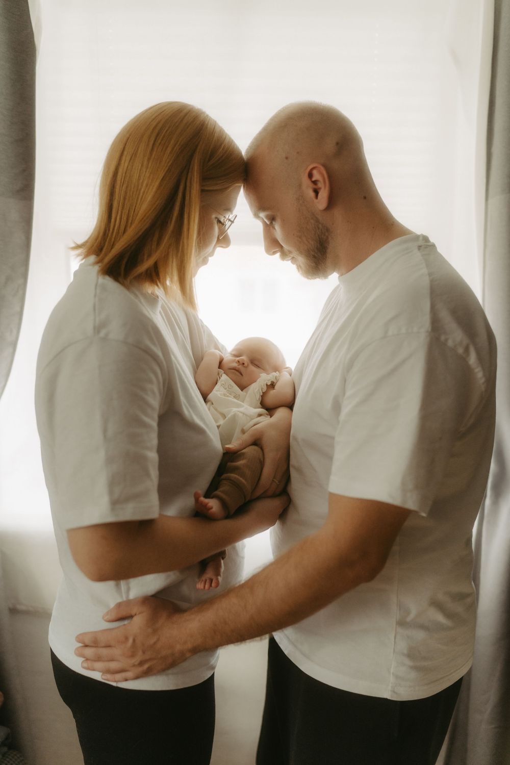 New parents in white t-shirts tenderly hold their newborn baby together near a bright window.