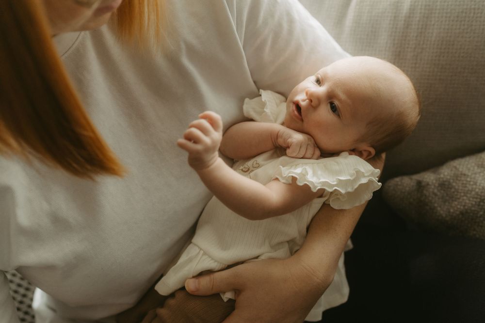 A mother in a white shirt gently cradles her newborn baby girl in a white ruffle dress while seated on a couch.